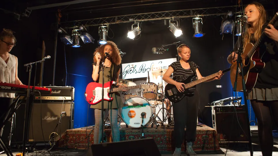 Band auf Bühne mit Instrumenten, bunte Beleuchtung, konzentriert spielend.