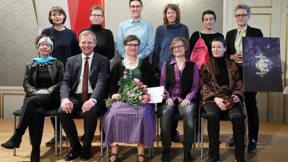 Menschen sitzen und stehen in zwei Reihen, einige halten Blumen und ein Poster.