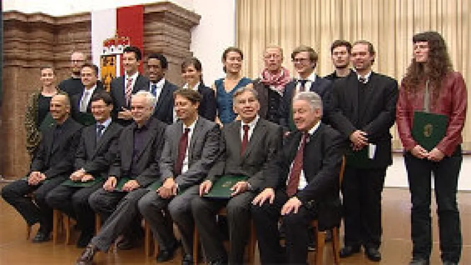 Gruppenfoto von 18 Personen in formeller Kleidung in einem Saal.