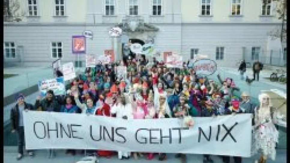 Menschenmenge mit Transparent "OHNE UNS GEHT NIX" vor einem Gebäude, fröhliche Stimmung.