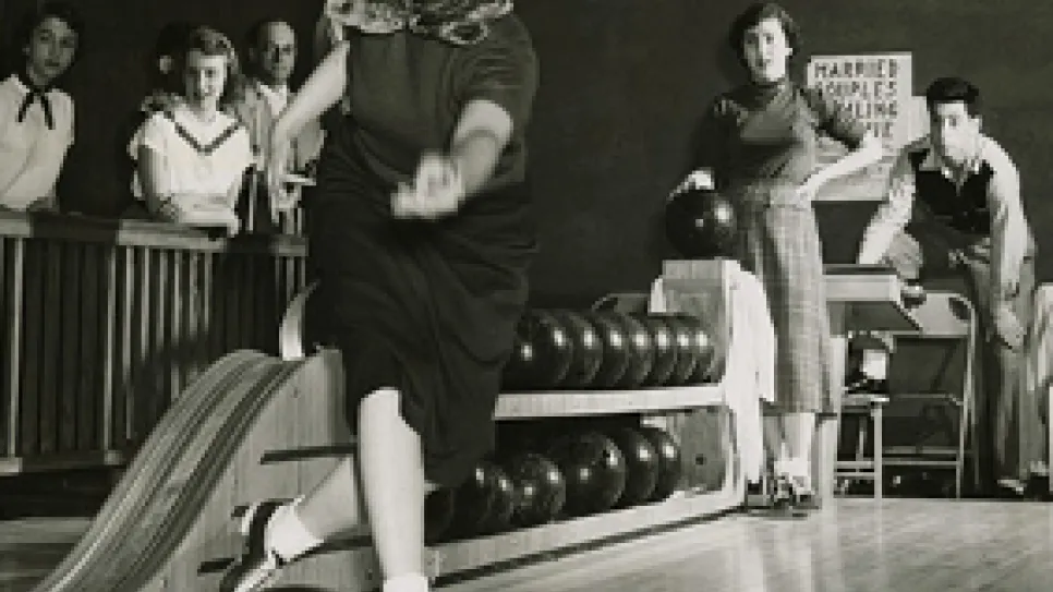 Frau in Bowlingbahn wirft Kugel, beobachtet von vier Personen. Schwarzweiß-Fotografie.