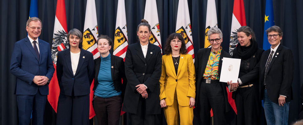 Eine Gruppe von acht Personen steht vor nationalen und regionalen Flaggen auf einer Bühne.