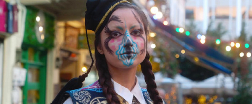 Person in Tracht mit bunter Gesichtsbemalung vor festlicher Kulisse.