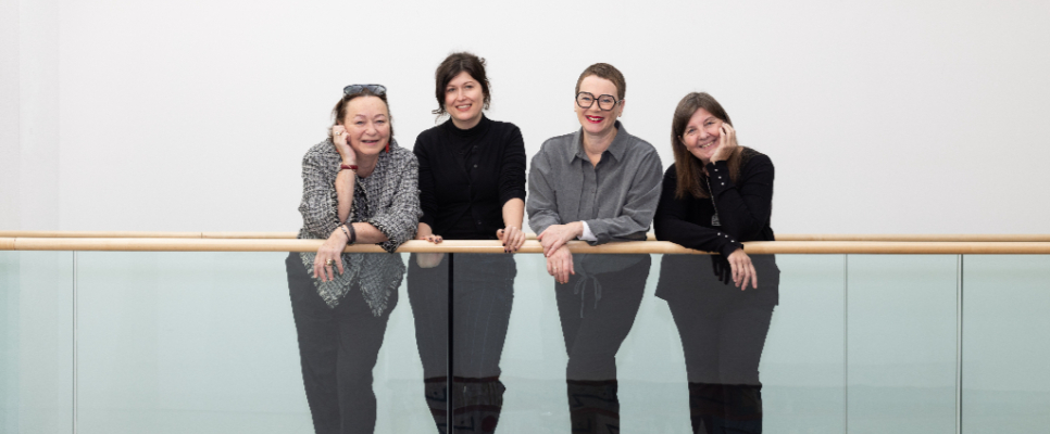 Vier Frauen lehnen lächelnd an einem Glasgeländer in einem modernen Gebäude.