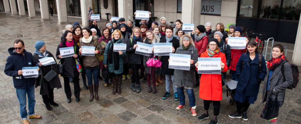 Gruppe von Menschen mit Schildern vor einem Gebäude, teils regenreich bekleidet.