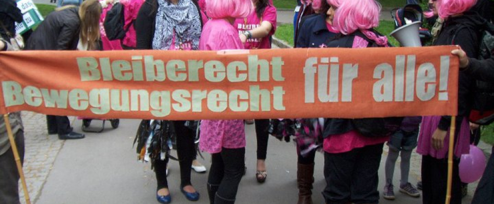 Personengruppe mit rosa Perücken, hält Banner: "Bleiberecht, Bewegungsrecht für alle!".