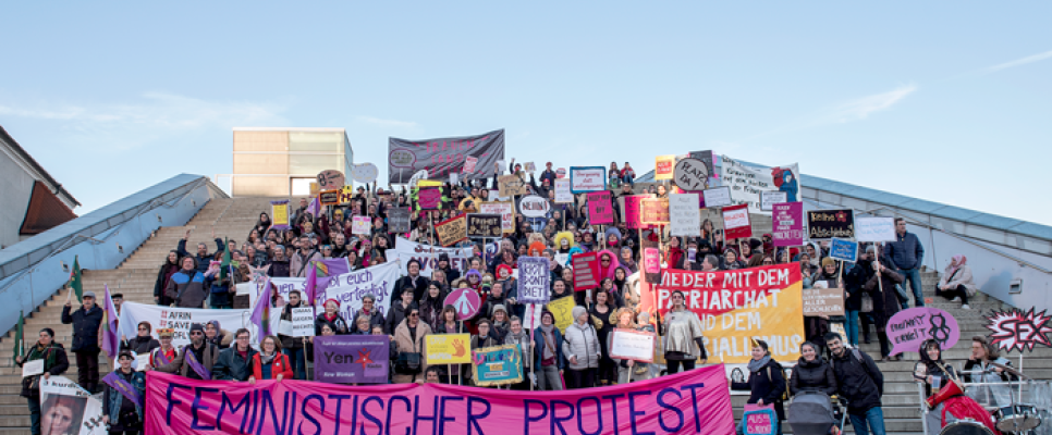 Protestierende Menschen auf Treppe mit Schildern und Banner, klarer Himmel.
