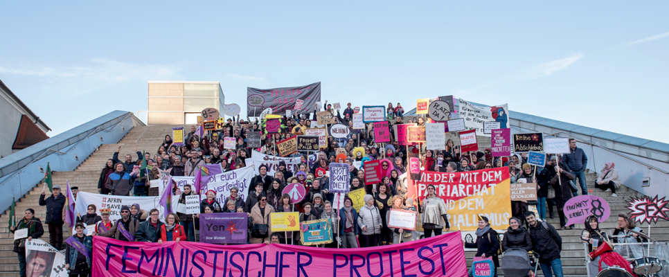 Menschenmenge auf Treppe mit Plakaten, Hauptbanner: "Feministischer Protest".