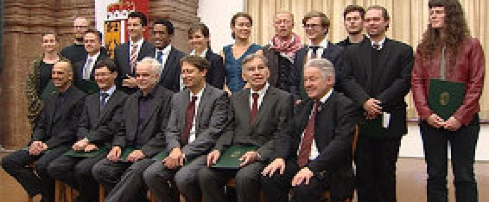 Gruppenfoto von 18 Personen in formeller Kleidung in einem Saal.