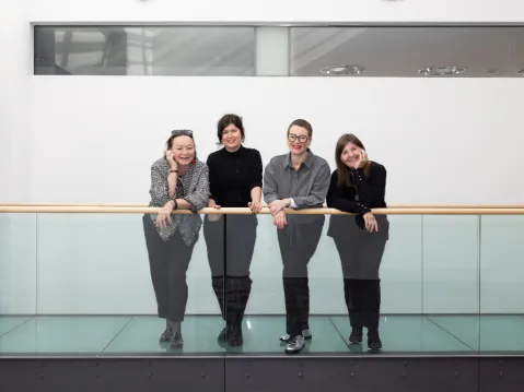 Vier Frauen lehnen lächelnd an einem Glasgeländer in einem modernen Gebäude.