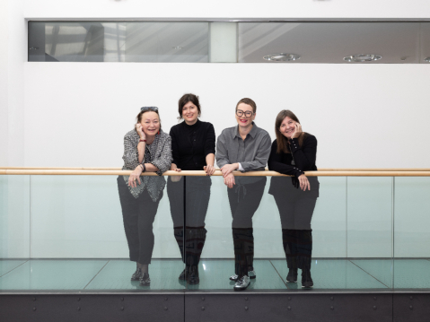 Vier Frauen lehnen lächelnd an einem Glasgeländer in einem modernen Gebäude.