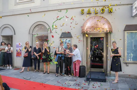 Menschen feiern vor einem Gebäude mit der Aufschrift "Salon", Konfetti in der Luft.