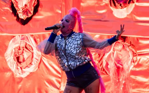 Person in Spiegeljacke und pinker Maske singt vor rotem Hintergrund.