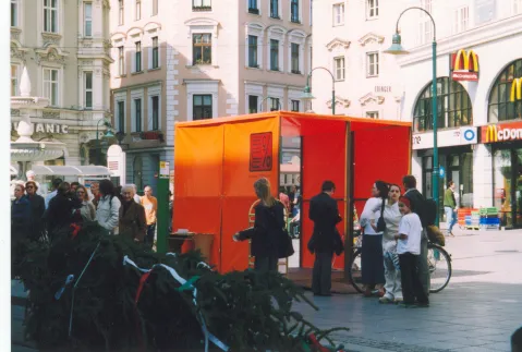 Rote Informationshütte auf belebtem Stadtplatz. Menschen stehen davor und unterhalten sich.