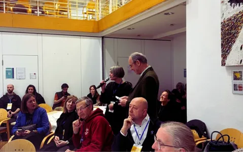 Versammlung in Konferenzraum, Person spricht in Mikrofon, Zuhörer aufmerksam.