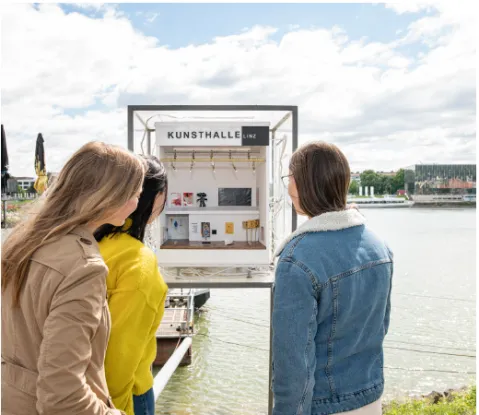 Drei Frauen betrachten Ausstellungsbox am Seeufer, bewölkter Himmel.