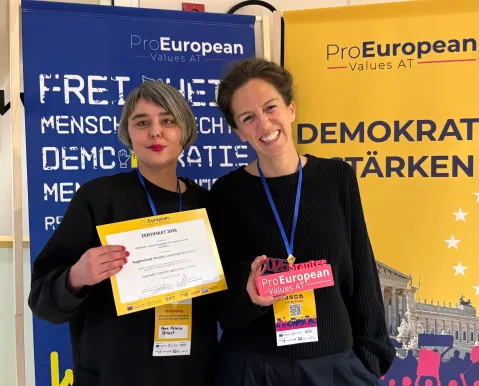 Zwei Frauen lächeln mit Zertifikat und Ausweis vor ProEuropean Plakaten.
