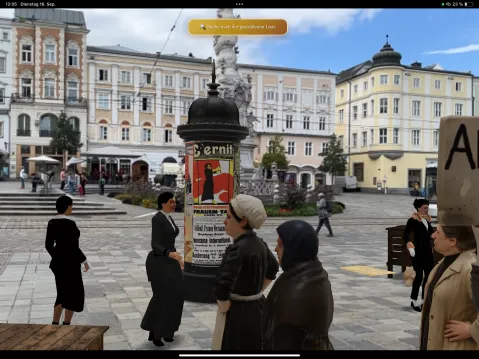 Menschen in historischer Kleidung versammeln sich um eine Litfaßsäule auf einem Stadtplatz.