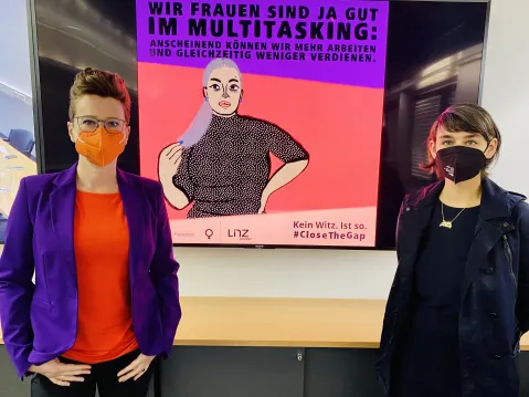 Zwei Frauen mit Masken vor feministischem Plakat zur Gender Pay Gap.