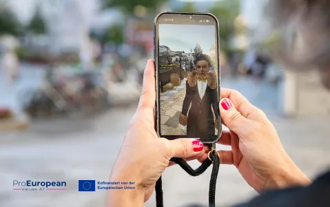 Person hält Smartphone mit Selfie-Kamera, Fokus auf Bildschirm, draußen. ProEuropean Logo sichtbar.