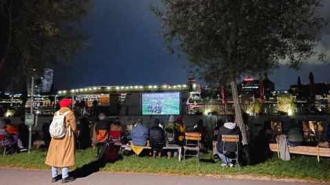 Menschen sehen abends draußen einen Film auf einer Leinwand, Umrisse der Stadt im Hintergrund.