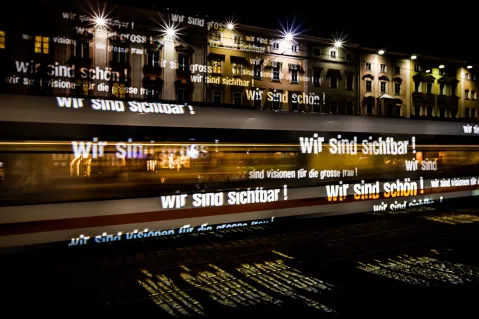 Lichter mit "Wir sind sichtbar!" auf vorbeifahrender Tram bei Nacht.