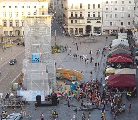 Menschenmenge um eine verhüllte Statue, bunte Schirme, helle Gebäude im Hintergrund.
