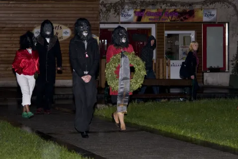 Menschen in Kostümen mit Masken und einem Kranz gehen einen Gehweg entlang, Nachtaufnahme.