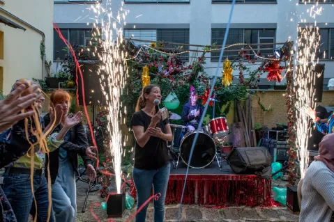 Eine Frau mit Mikrofon vor Bühne mit Band und bunten Lichtern, umgeben von Feiernden.