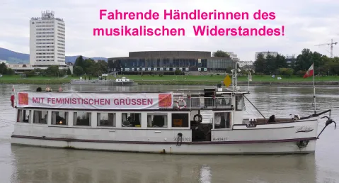 Ein Boot auf einem Fluss mit einem Banner an Deck, städtische Kulisse im Hintergrund.