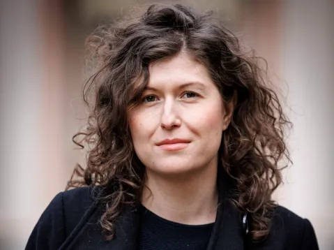 Eine Frau mit lockigem Haar, neutraler Ausdruck, draußen.