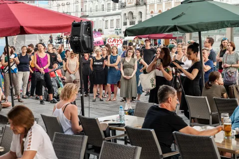 Menschenmenge auf einem Platz, einige halten Schilder, im Vordergrund Personen an Tischen.