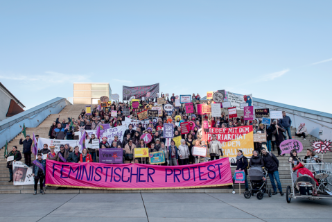 Protestierende Menschen auf Treppe mit Schildern und Banner, klarer Himmel.