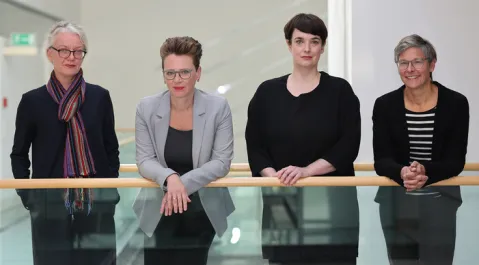 Vier Frauen stehen nebeneinander an einem Geländer und lächeln.