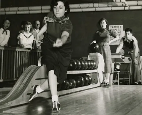 Frau in Bowlingbahn wirft Kugel, beobachtet von vier Personen. Schwarzweiß-Fotografie.
