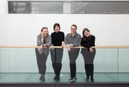 Vier Frauen lehnen lächelnd an einem Glasgeländer in einem modernen Gebäude.