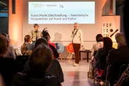 Zwei Frauen auf Bühne, halten Vortrag vor Publikum, feministische Themen, beleuchteter Raum.