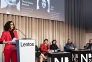 Rednerin im roten Anzug am Podium, fünf Frauen sitzen, minimalistischer Raum.