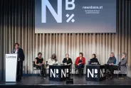 Podiumsdiskussion mit sieben Frauen auf einer Bühne, Logo "NB" im Hintergrund.