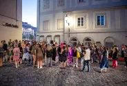 Menschenansammlung auf gepflasterter Straße bei Dämmerung, diskutierend vor beleuchtetem Gebäude.