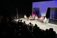 Podiumsdiskussion mit sieben Personen auf einer Bühne, Publikum im Vordergrund.