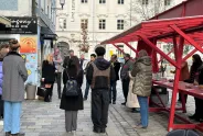 Menschenmenge bei einem roten Stand auf Kopfsteinpflasterstraße, städtische Umgebung.