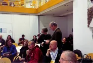 Versammlung in Konferenzraum, Person spricht in Mikrofon, Zuhörer aufmerksam.