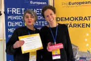 Zwei Frauen lächeln mit Zertifikat und Ausweis vor ProEuropean Plakaten.