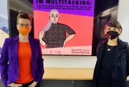 Zwei Frauen mit Masken vor feministischem Plakat zur Gender Pay Gap.