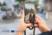 Person hält Smartphone mit Selfie-Kamera, Fokus auf Bildschirm, draußen. ProEuropean Logo sichtbar.
