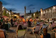 Menschen sitzen auf Liegestühlen bei Abenddämmerung auf einem belebten Platz.