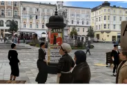 Frauen in historischer Kleidung auf einem Platz mit Plakatsäule, umgeben von alten Gebäuden.