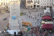 Menschenmenge um eine verhüllte Statue, bunte Schirme, helle Gebäude im Hintergrund.