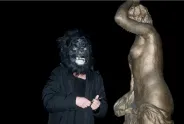 Person mit Gorilla-Maske neben goldener Statue bei Nacht.